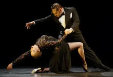 Male dancer in a tuxedo supports a female dancer in a black sequined dress as she leans back in a dramatic ballroom pose on a dark stage.