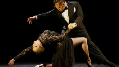 Male dancer in a tuxedo supports a female dancer in a black sequined dress as she leans back in a dramatic ballroom pose on a dark stage.