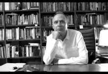 Man in a white shirt sits at a desk in a library, resting his chin on his hand.