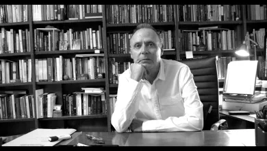 Man in a white shirt sits at a desk in a library, resting his chin on his hand.