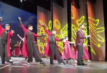 Group of dancers in red and black outfits performing a choreographed routine on a stage with yellow abstract light projections in the background.