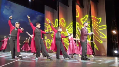 Group of dancers in red and black outfits performing a choreographed routine on a stage with yellow abstract light projections in the background.