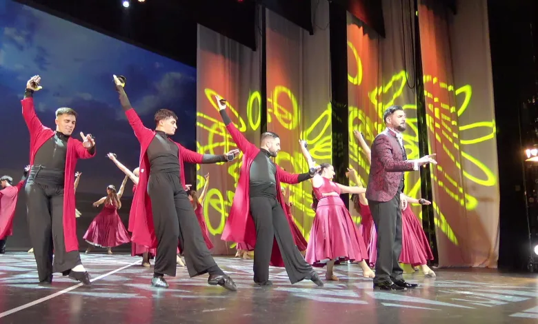 Group of dancers in red and black outfits performing a choreographed routine on a stage with yellow abstract light projections in the background.