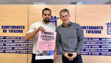 Two Men Smile for the Camera in Front of a Tarazona City Hall Backdrop the Man on the Left Holds a Pink Event Poster Tarazona Acoge babeando Ternasco El 6 De Junio Zaragozala Qué Hacer En Zaragoza