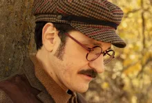 Man wearing a brown tweed jacket and plaid newsboy cap leaning against a tree in a sunlit autumn forest.