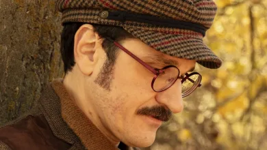 Man wearing a brown tweed jacket and plaid newsboy cap leaning against a tree in a sunlit autumn forest.