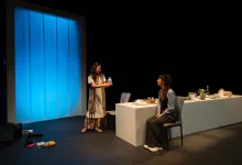 Two women on a theater stage; one standing hands a jar to the seated woman at a white desk with props nearby, blue backdrop left.
