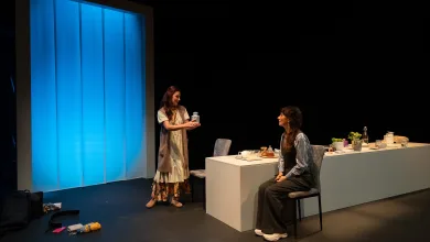 Two women on a theater stage; one standing hands a jar to the seated woman at a white desk with props nearby, blue backdrop left.