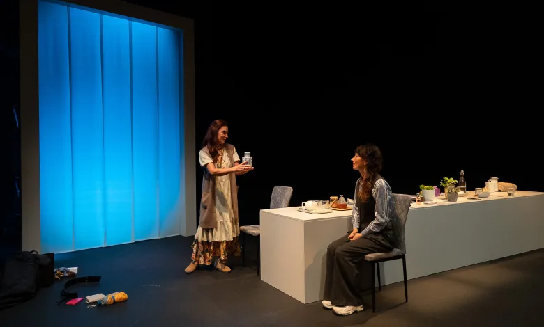 Two women on a theater stage; one standing hands a jar to the seated woman at a white desk with props nearby, blue backdrop left.