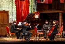 Four musicians perform on a stage—pianist, violinist, percussionist, and cellist—against red curtains and a grand piano in a vintage concert hall.