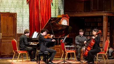 Four musicians perform on a stage—pianist, violinist, percussionist, and cellist—against red curtains and a grand piano in a vintage concert hall.
