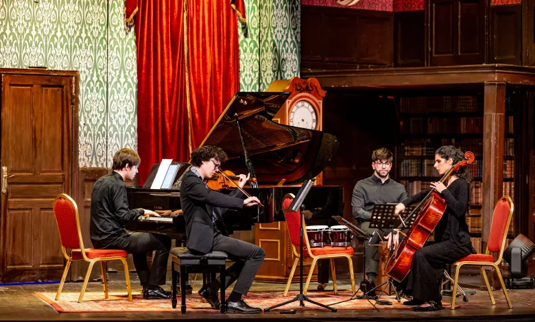 Four musicians perform on a stage—pianist, violinist, percussionist, and cellist—against red curtains and a grand piano in a vintage concert hall.