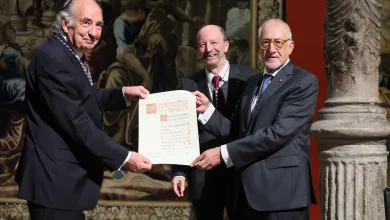 Three men in suits exchange a certificate during a formal award ceremony, with an ornate mural in the background.