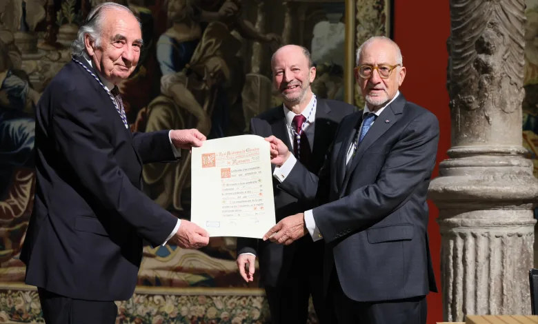 Three men in suits exchange a certificate during a formal award ceremony, with an ornate mural in the background.