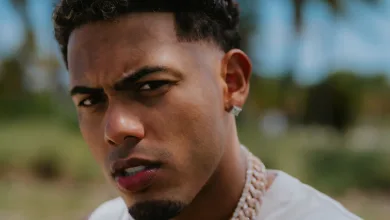 Close-up portrait of a young man with short curly hair, wearing a diamond stud earring and a thick chain necklace, squinting at the camera outdoors.