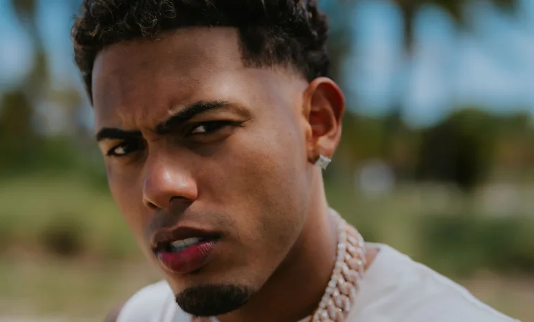 Close-up portrait of a young man with short curly hair, wearing a diamond stud earring and a thick chain necklace, squinting at the camera outdoors.