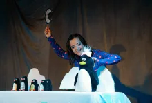 Performer in a blue pajama top raises a hat on a stage, with a large penguin puppet and a row of penguin figures on a table nearby