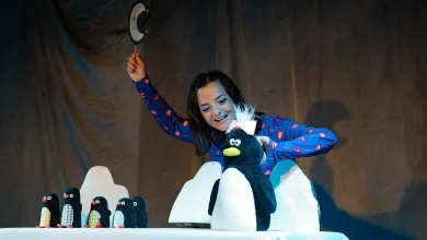 Performer in a blue pajama top raises a hat on a stage, with a large penguin puppet and a row of penguin figures on a table nearby