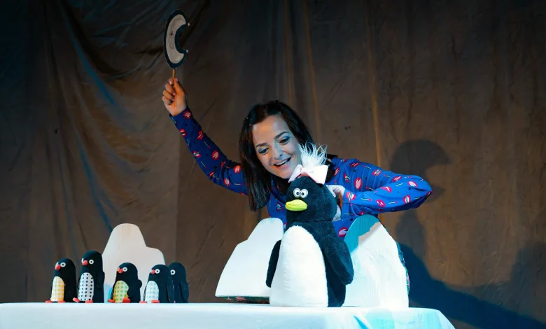 Performer in a blue pajama top raises a hat on a stage, with a large penguin puppet and a row of penguin figures on a table nearby