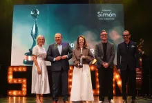 Five people stand on a stage smiling; the center woman holds a trophy during an awards ceremony with a large screen behind them and illuminated letters on stage.