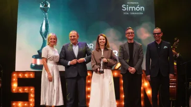 Five people stand on a stage smiling; the center woman holds a trophy during an awards ceremony with a large screen behind them and illuminated letters on stage.