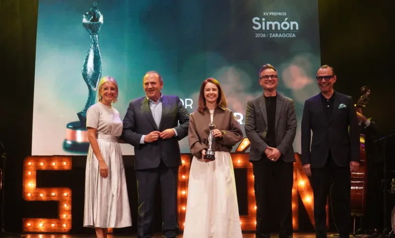 Five people stand on a stage smiling; the center woman holds a trophy during an awards ceremony with a large screen behind them and illuminated letters on stage.