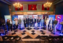 Group of professionals posing in a marble-floored venue with sponsor banners and chandeliers overhead.