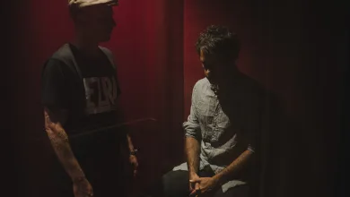 Two men stand opposite each other in a dim, red-lit room; one wears a cap, the other sits and looks down.