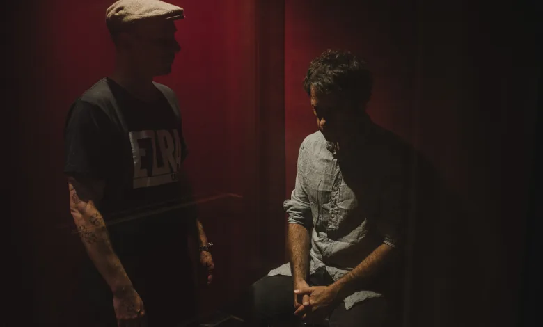 Two men stand opposite each other in a dim, red-lit room; one wears a cap, the other sits and looks down.