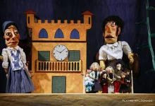 Puppet show scene with a tall orange building backdrop and a clock, flanked by two large puppets, with many small puppets gathered in front.