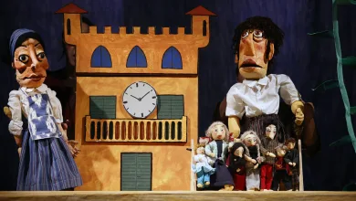 Puppet show scene with a tall orange building backdrop and a clock, flanked by two large puppets, with many small puppets gathered in front.