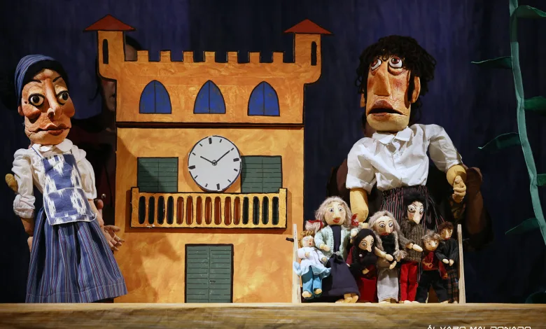 Puppet show scene with a tall orange building backdrop and a clock, flanked by two large puppets, with many small puppets gathered in front.