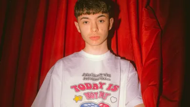 Young person standing in front of red curtains, wearing a white T-shirt with bold dice graphics and the text 'TODAY IS NOT MY'.