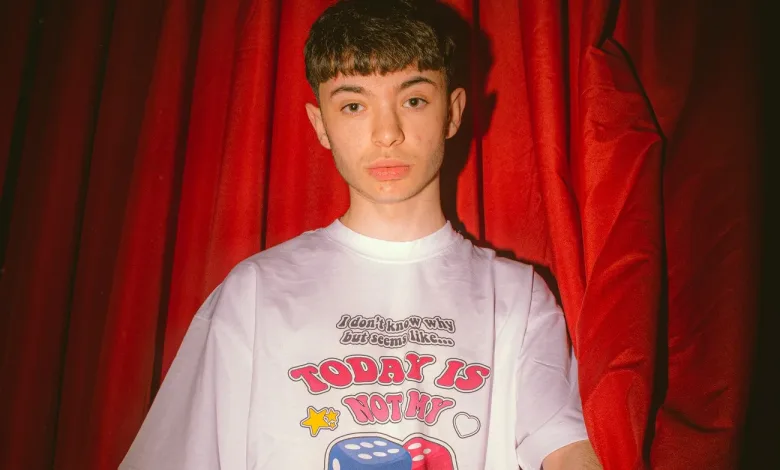 Young person standing in front of red curtains, wearing a white T-shirt with bold dice graphics and the text 'TODAY IS NOT MY'.