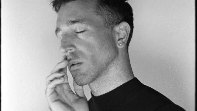 Close-up of a man with eyes closed, hands pressed together near his lips, wearing a dark sweater.