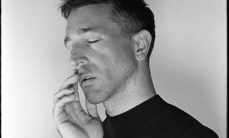 Close-up of a man with eyes closed, hands pressed together near his lips, wearing a dark sweater.
