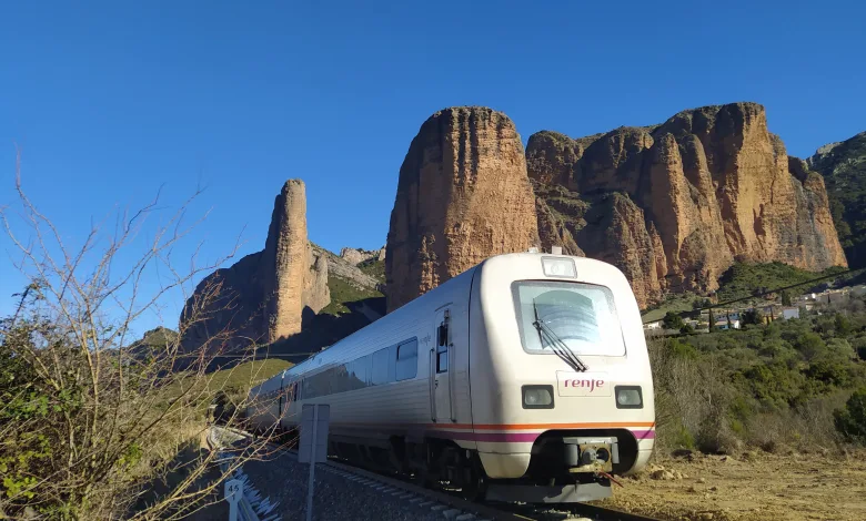 White Renfe train on railroad tracks beside rugged red cliffs under a clear blue sky, with dry vegetation in the foreground.