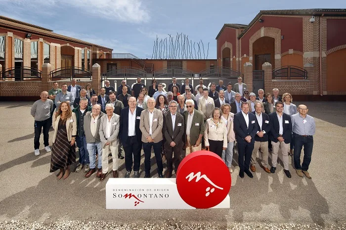 Group of about 40 people posing outdoors for a formal photo in front of a building, with a white banner and red circular logo reading Somontano.