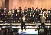 A choir of about twenty people in black shirts performs on a stage, with a conductor facing them from the front and an audience watching theater-style seating nearby.
