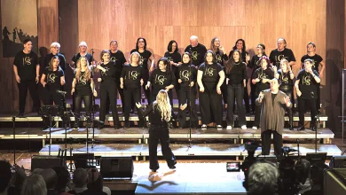 A choir of about twenty people in black shirts performs on a stage, with a conductor facing them from the front and an audience watching theater-style seating nearby.
