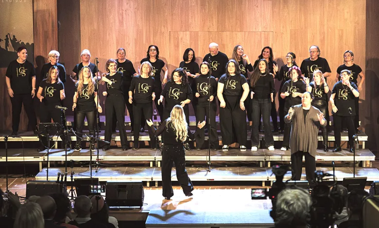 A choir of about twenty people in black shirts performs on a stage, with a conductor facing them from the front and an audience watching theater-style seating nearby.