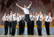 Group of men in white shirts and black ties on a stage, one man jumps mid-air as others cheer around him on a podium backdrop.