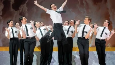 Group of men in white shirts and black ties on a stage, one man jumps mid-air as others cheer around him on a podium backdrop.