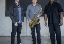 Three men pose in front of a concrete wall; center man holds a saxophone, wearing a gray shirt and necklace.