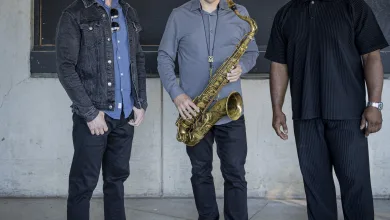 Three men pose in front of a concrete wall; center man holds a saxophone, wearing a gray shirt and necklace.