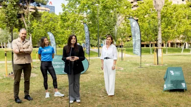 Four adults stand in a sunny park for an outdoor event, with a speaker at a microphone and tall banners in the background.