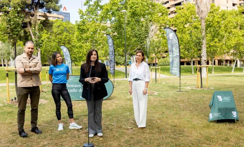 Four adults stand in a sunny park for an outdoor event, with a speaker at a microphone and tall banners in the background.