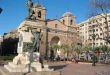 Bronze statue on a pedestal in a sunny plaza with a historic arched church in the background and palm trees nearby, pedestrians strolling nearby.