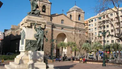 Bronze statue on a pedestal in a sunny plaza with a historic arched church in the background and palm trees nearby, pedestrians strolling nearby.