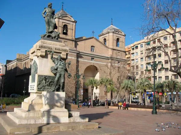 Bronze statue on a pedestal in a sunny plaza with a historic arched church in the background and palm trees nearby, pedestrians strolling nearby.